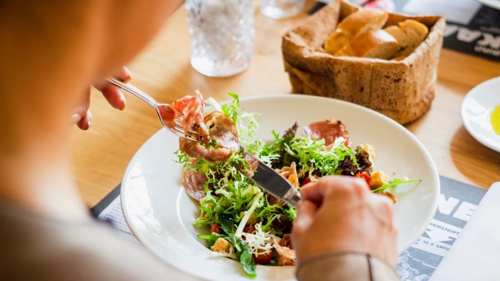 A person eating a salad with cured meat and croutons from a white plate
